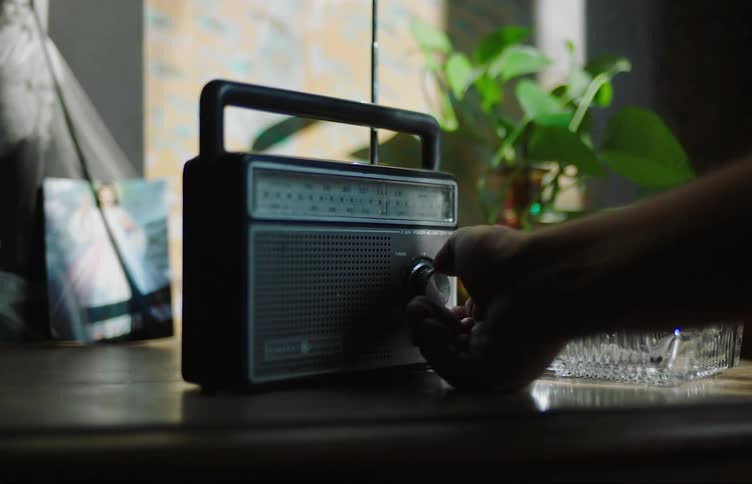 A hand reaches towards an old radio next to an ashtray with a lit cigarette on it and spins a dial