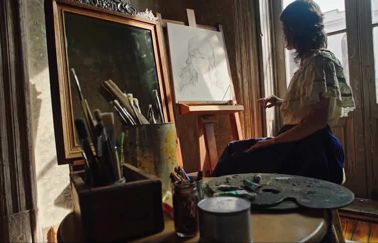 A person is painting a self portrait on a canvas by a window in a room filled with art supplies