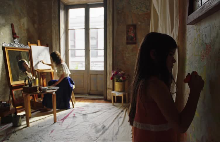 A girl draws on the wall while a woman paints on canvas behind her