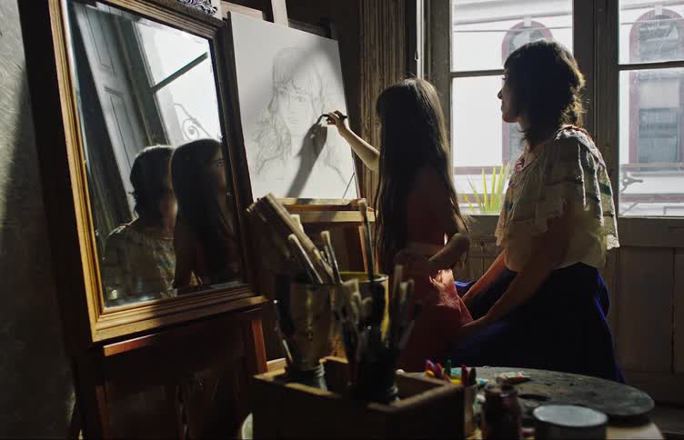 Mother sitting in a chair with her daughter as she begins to draw the features of a face on a white canvas