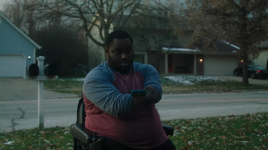 Man with no hands and no legs sitting in an automated chair playing on his cell phone while on a sidewalk in a residence
