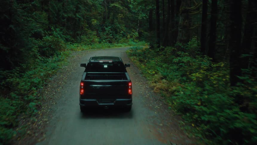 A black pickup truck drives on a narrow road through a forest