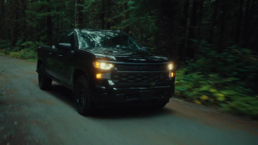 A black pickup truck with headlights on driving on a road with many trees around it