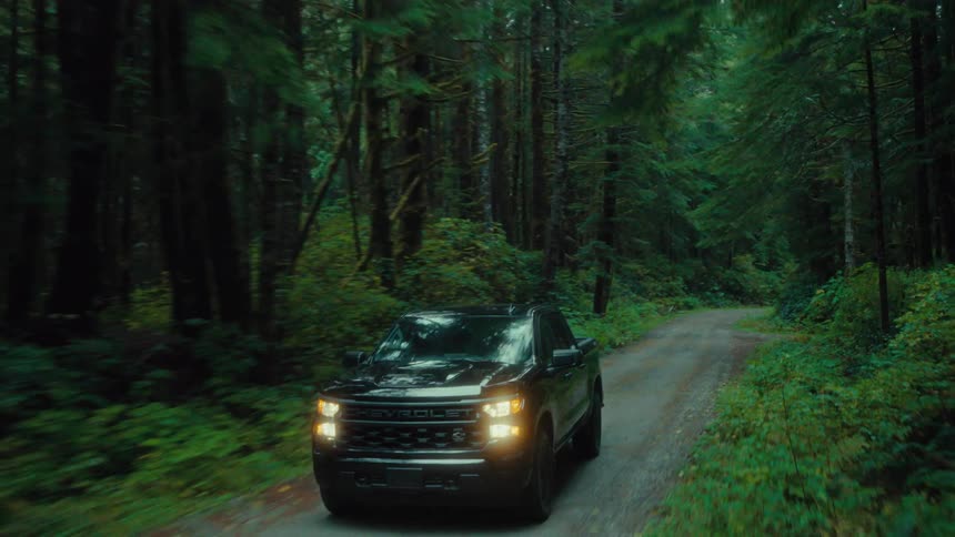 Black pickup truck with headlights on traveling along a narrow road surrounded by forest