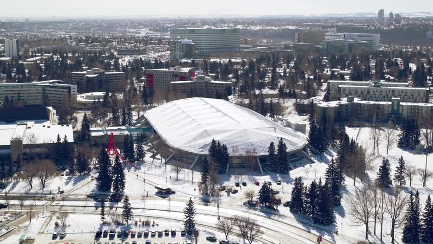 Aerial view of an indoor skating oval and surrounding residential and urban buildings in a city covered in snow