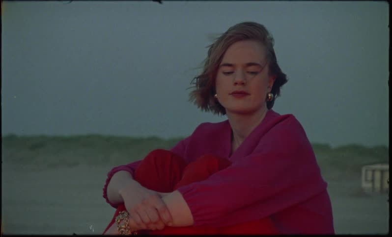 Vintage footage of a woman in red sitting near flames in the desert on a windy day