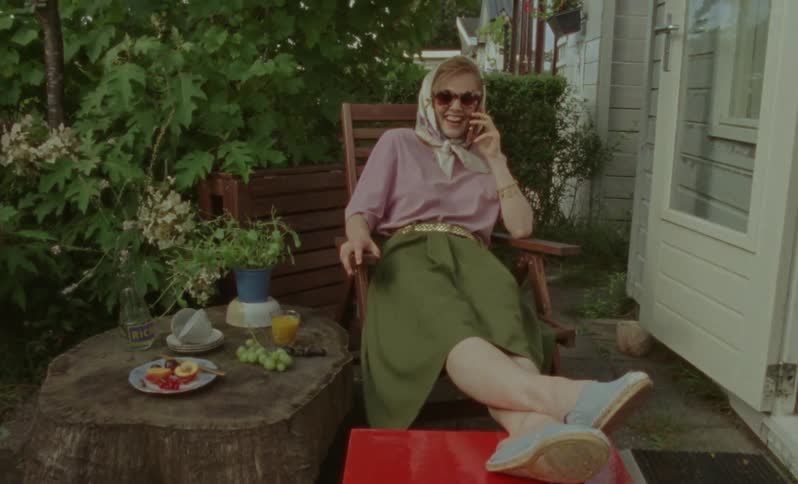 A woman in a dress and sunglasses reclines outdoors while talking on a cell phone
