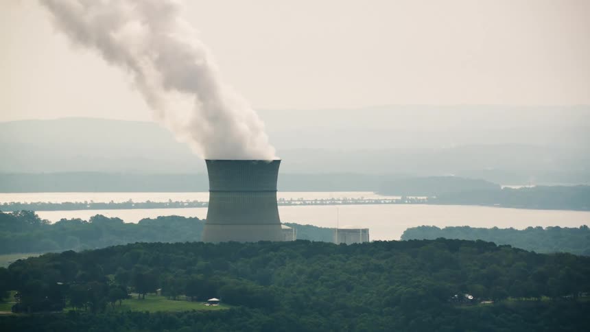 Aerial panorama of a large chimney in a factory releasing smoke amid forested hills and the sea