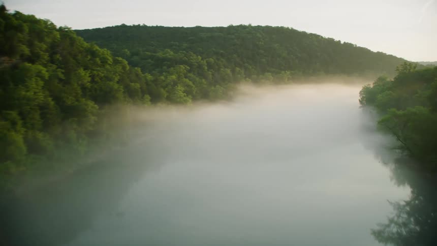 Fog partially obscures river reflecting forested terrain near mountain with a building