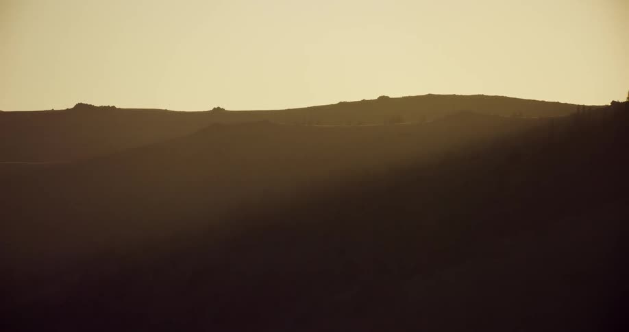 Amber sunlight shines from red sky onto conifer forest on hills