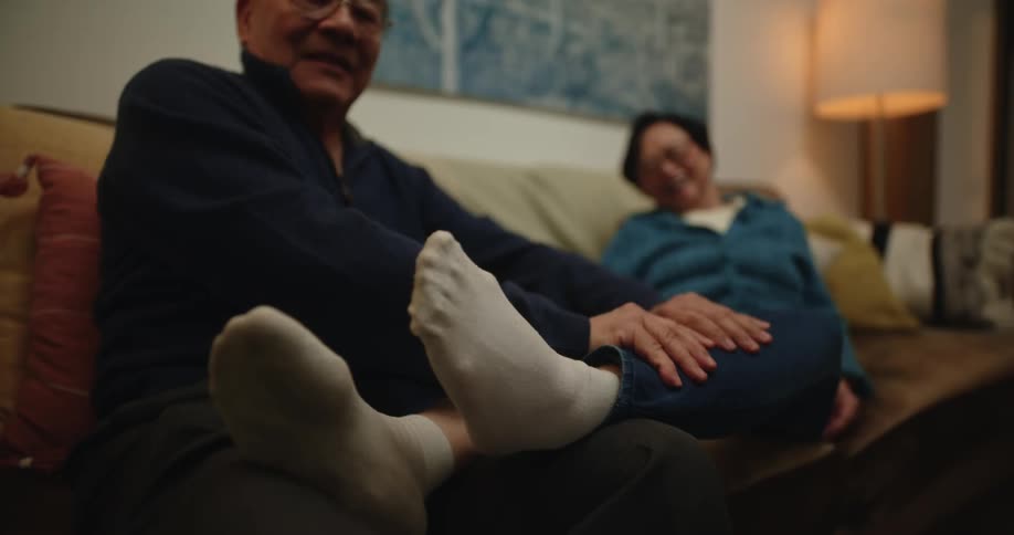 Elderly couple sitting on sofa with wifes legs being massaged by husband