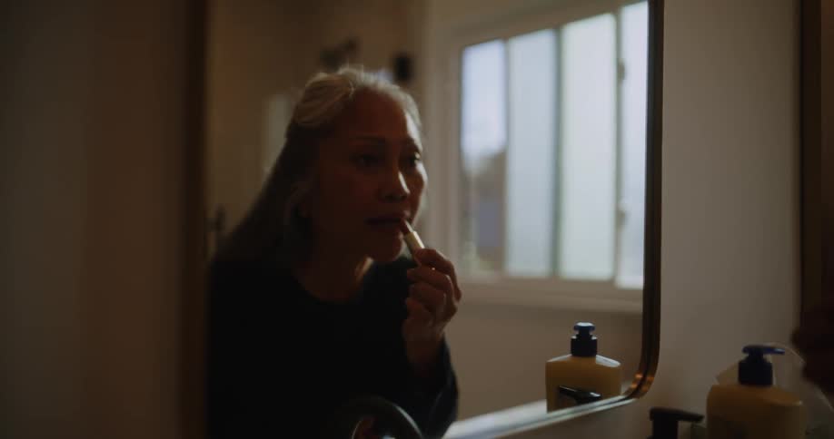 Elderly woman applying lipstick in front of a mirror in the bathroom