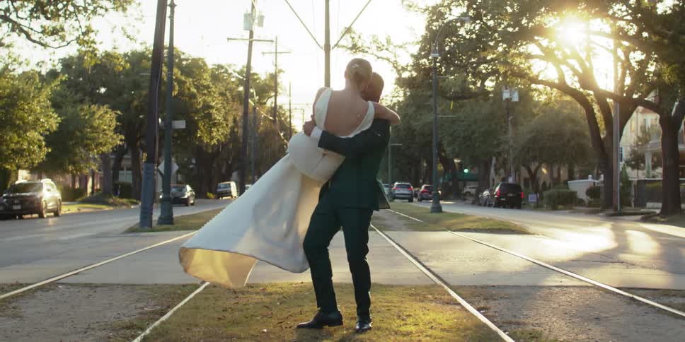 Man in suit holding his bride in a long white dress on his lap and spinning her around as they stand on a sidewalk in the middle of an urban street