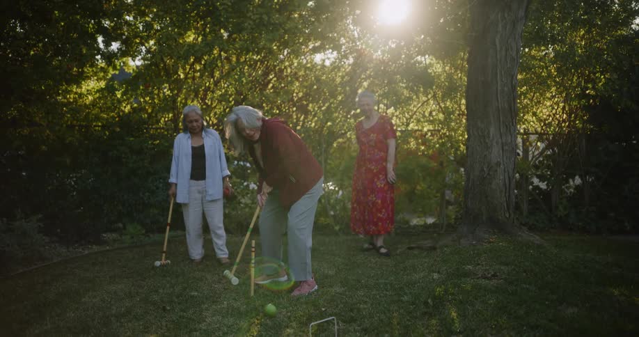 Three older female friends play croquet on lawn near fence on a sunny day