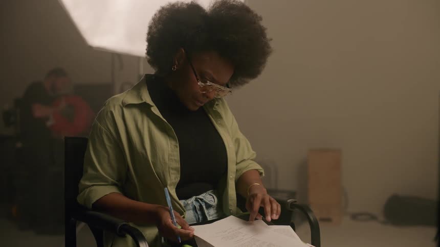 Woman sitting in a chair taking notes on a sheet of paper while two other people in the background work in a studio