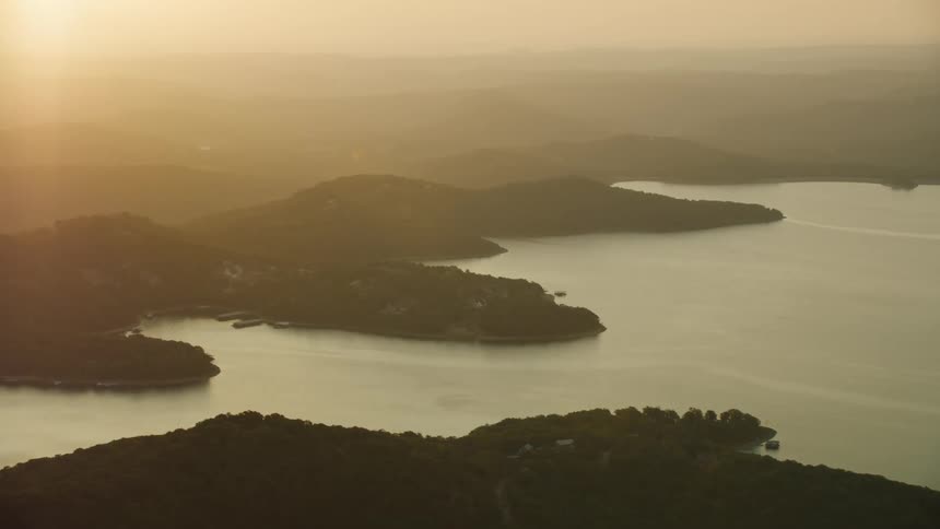 Aerial view of a lake surrounded by lush green vegetation and rolling hills covered in fog with the glare of a low sun in the background