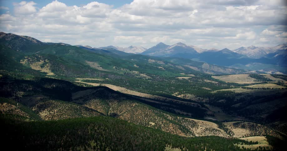 Aerial view of a valley with deep green vegetation scattered on tan soil and contrasting white clouds and blue mountains in the background