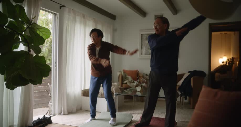 Two elderly asian people stretch together in a living room