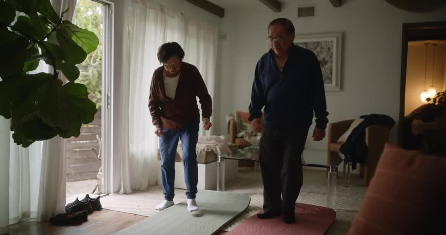 Elderly lady on a mat and next to her husband who imitates her in the tai chi movements she performs