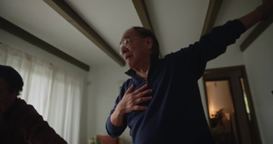 Elderly woman next to her husband both standing in the living room and him imitating the arm and body movements that his wife makes
