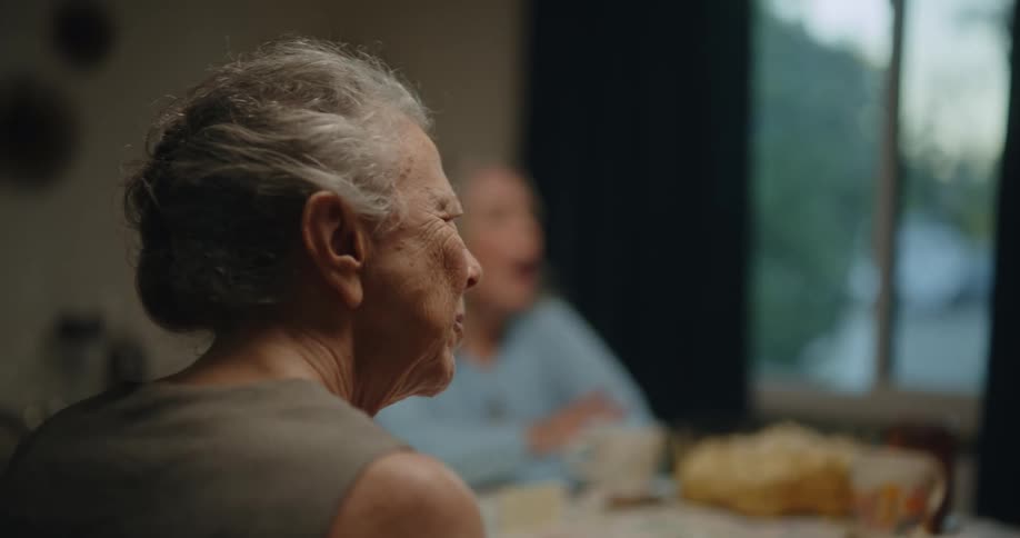 Three elderly lady friends sitting at a table talking to each other eating and playing dominoes