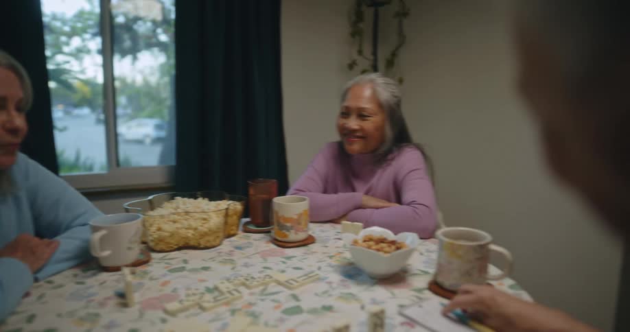 Three older women sitting at a round table eating and playing dominoes indoors