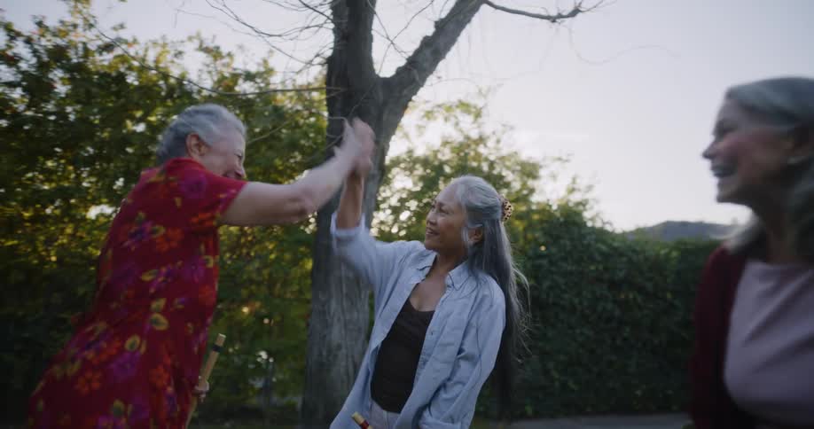Three retirement aged woman with silver hair playing croquette and high fiving in yard as the sunsets behind the trees in the background