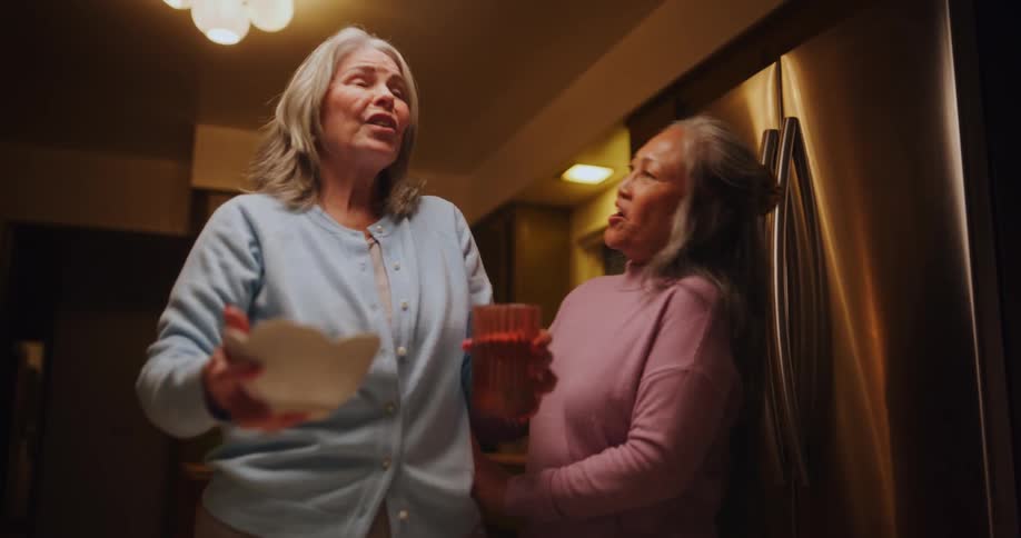 Elderly woman removing a glass of water from the table and a bowl being startled when her friend arrives behind her