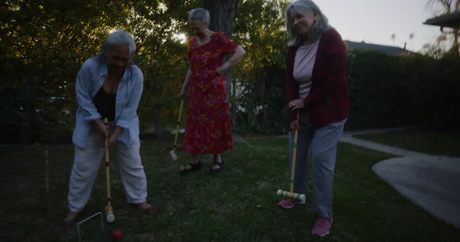 Three elderly friends standing in the backyard playing bat with one of them cheating and kicking the ball with her legs