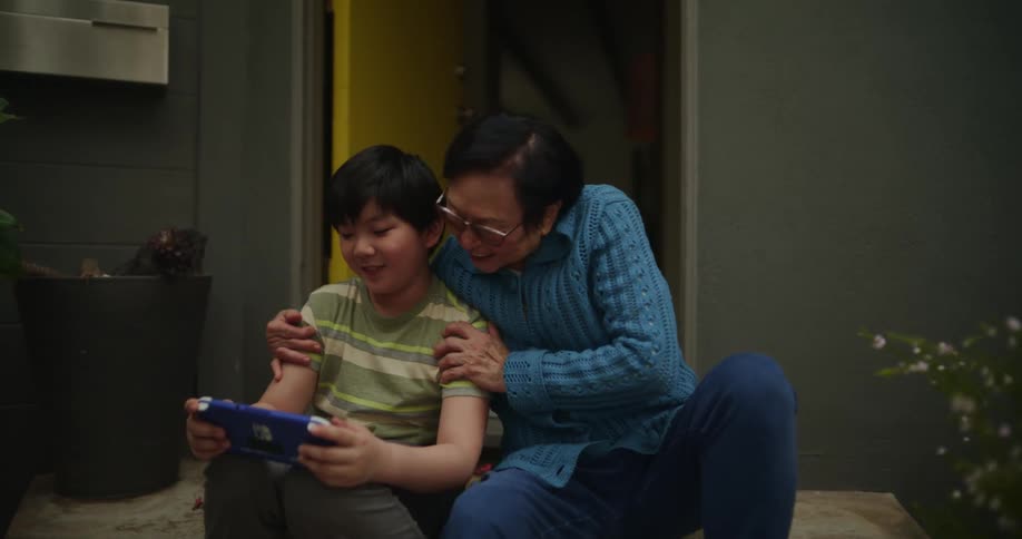 Grandfather sitting in the backyard with his grandson and they have fun while the grandson is playing on a portable game and they clap their hands together happily