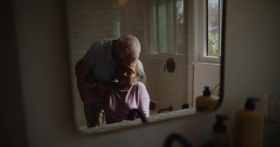 Elderly man combing his wifes hair who is sitting on a chair in the bathroom and then kissing her head