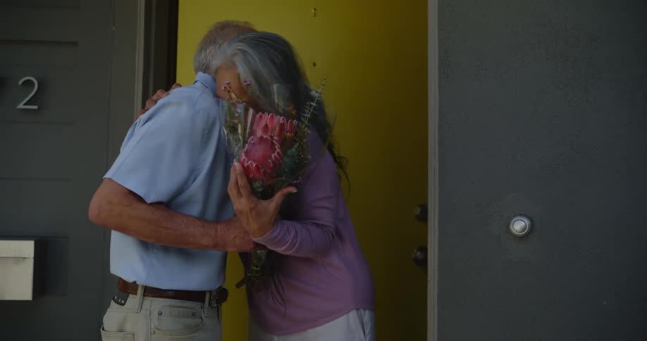 The lady opens the door and her romantic partner hands her a bouquet of roses they hug and then enter the house