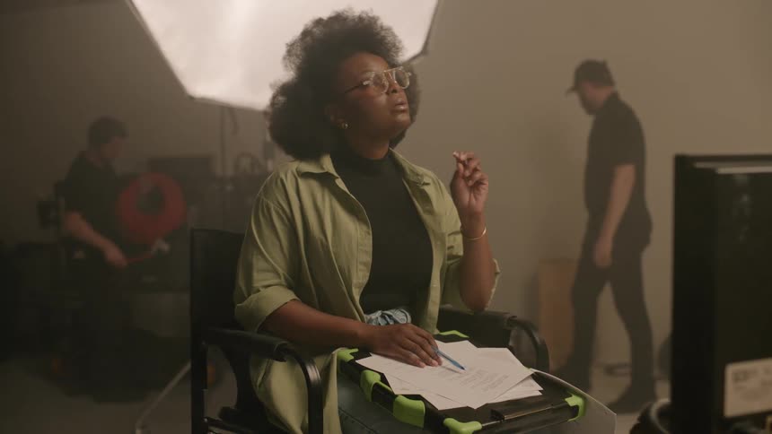 Woman sitting in a chair inside a studio taking notes on a script and in the background two men working with cables