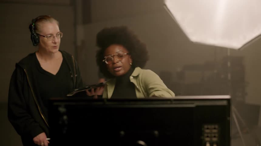 A director in a studio watches a scene on a monitor and then gives instructions to her assistant and the crew