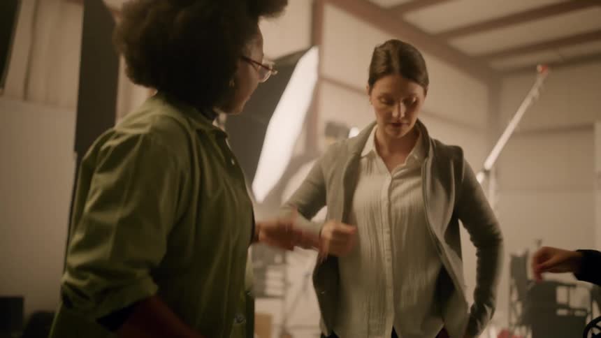 Two women assisting another woman who is getting dressed in a gray sweater inside a recording studio