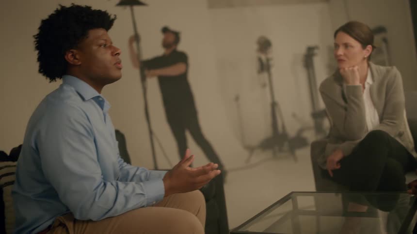 Three people sitting in armchairs facing each other talking while a man in the background turns on a studio light in the room