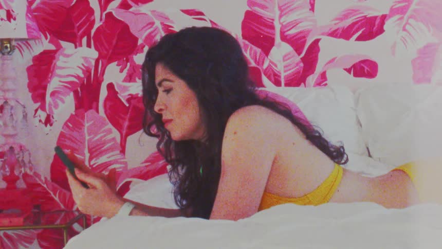 Woman wearing a yellow swimsuit checks her phone on the bed