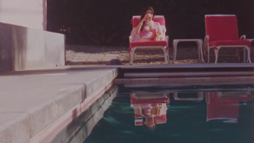 Woman in a pink swimsuit lying on a chair drinking wine facing a swimming pool