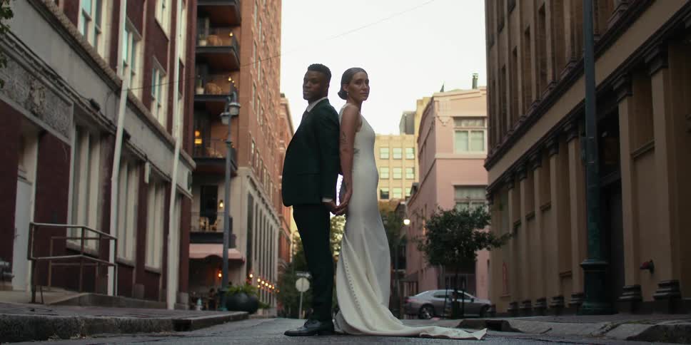Wedding couple in suit and long white dress holding hands and back to back on a street in the city