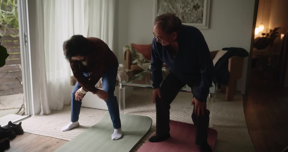 Elderly couple perform body stretches in which the husband imitates his wifes movements and they have fun