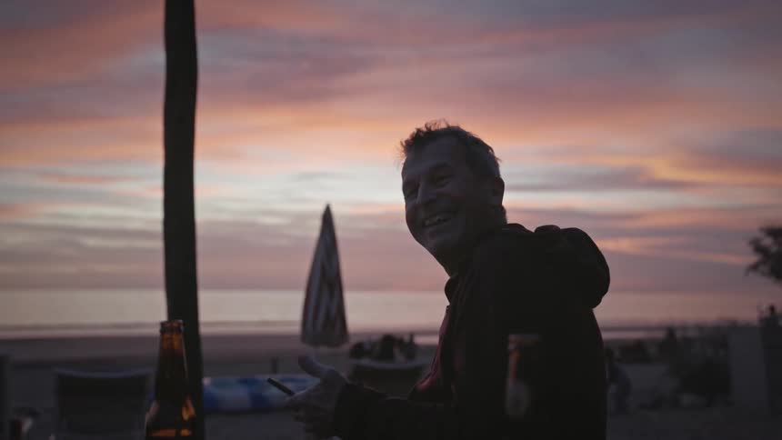 Man playing on his cell phone and laughing while sitting in front of the beach in a purple late afternoon sky