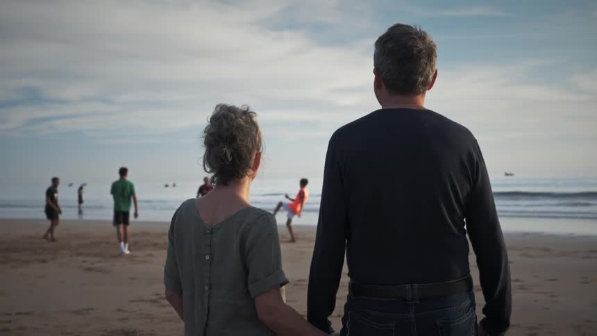 Man and woman holding hands near the beach watching people kick a ball