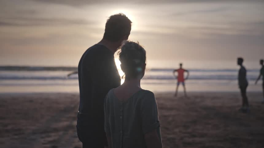 Husband and wife on the beach watching some people playing footvolley on a late afternoon with the sun on the horizon