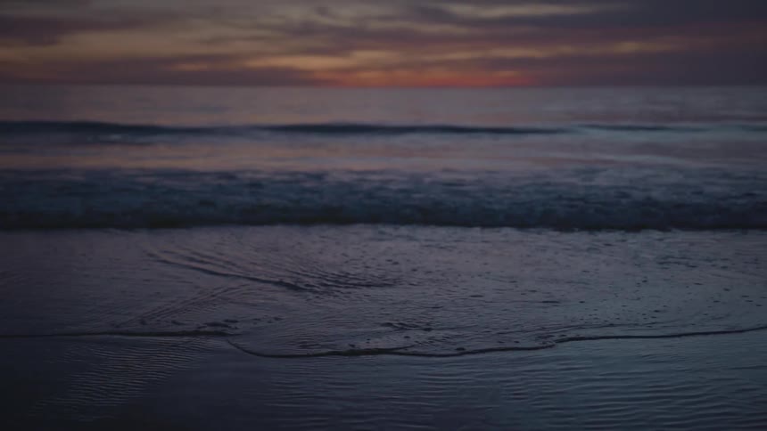 Ocean waves are crashing towards the coast of the beach at dusk