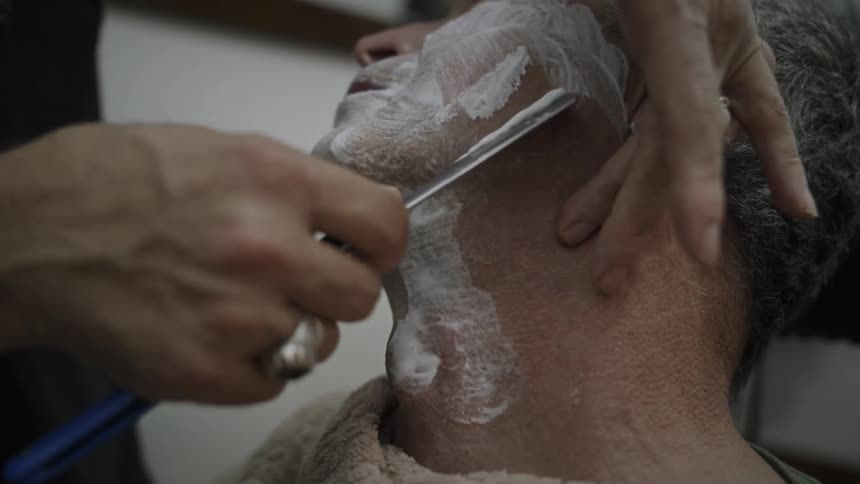 An elderly man leans his head back while having his face shaved