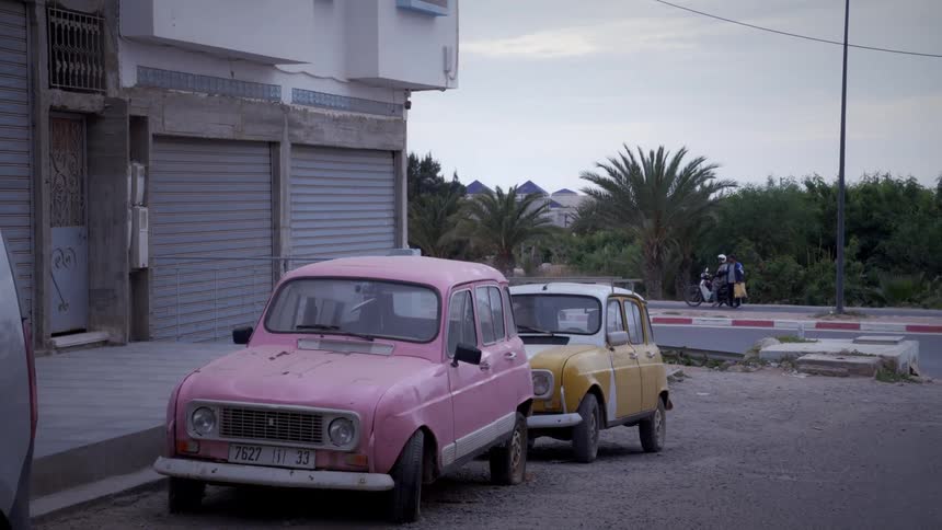 Two vintage cars parked on the street one pink and the other yellow and rusty