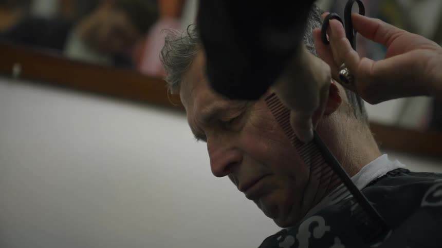 An elderly man sitting in a barber chair has his hair cut by a younger man