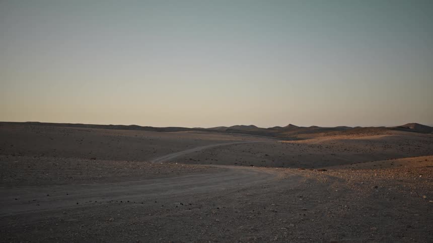 Vehicle trail curves across a sandy desert terrain at sunset