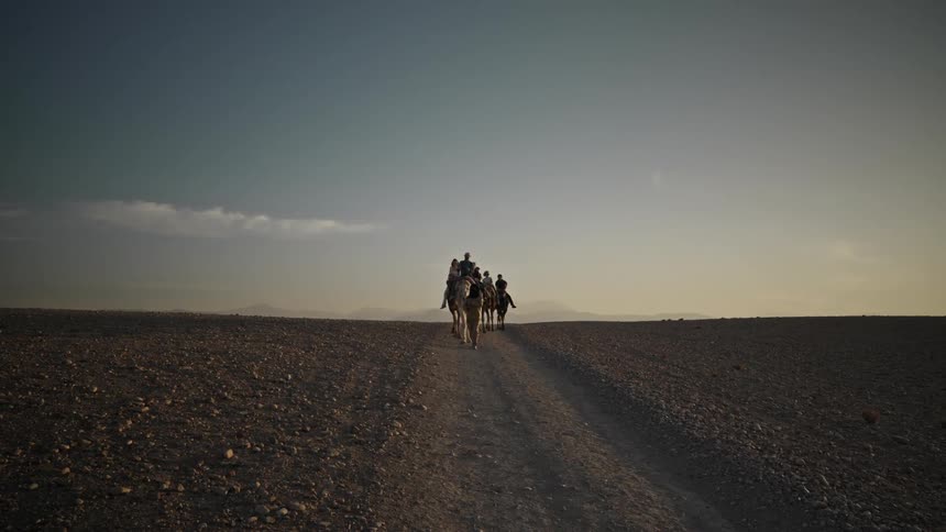 A group of people are riding camels along a dirt path in a vast desert