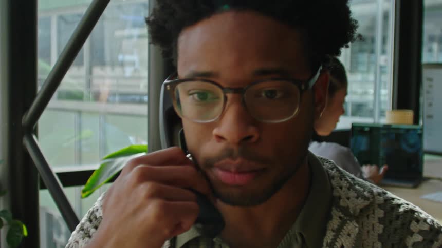 Man sitting in an office talking on the phone and in the background a woman working on the laptop